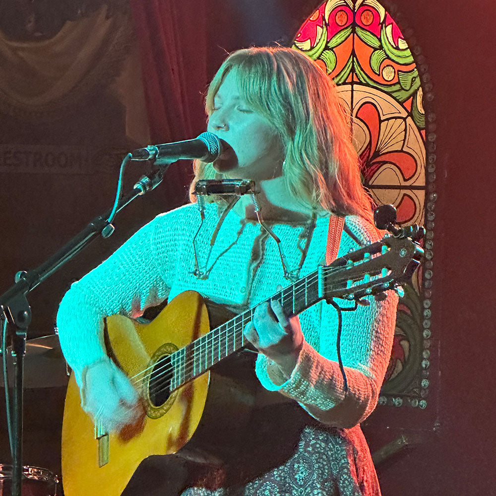 Person playing guitar and singing into a microphone with a colorful stained glass window in the background.